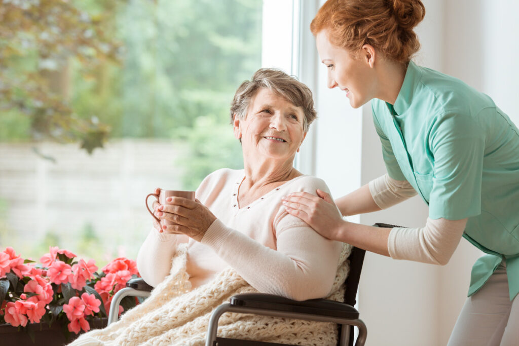 A caretaker touching the shoulders of an elderly woman holding a mug by a window in El Paso.