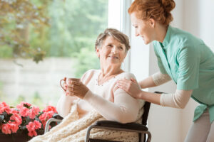 A caretaker touching the shoulders of an elderly woman holding a mug by a window in El Paso.